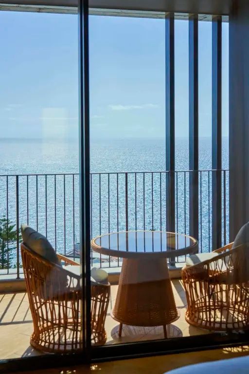 Blick vom Balkon durch bodentiefe Fenster auf das blaue Meer mit Tisch und zwei Stühlen.