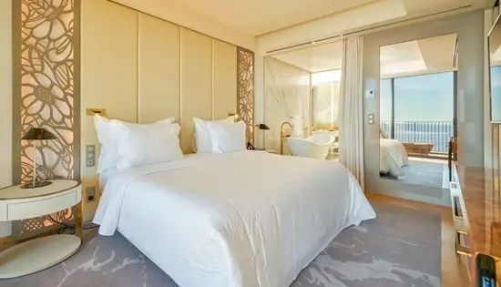 Doppelbett im Hotelzimmer mit hellen Kissen und Blick durch eine Glaswand ins Badezimmer.