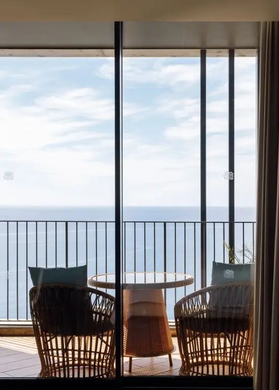 Blick von einem Balkon durch eine Glastür auf das offene Meer und zwei Stühle mit Tisch.