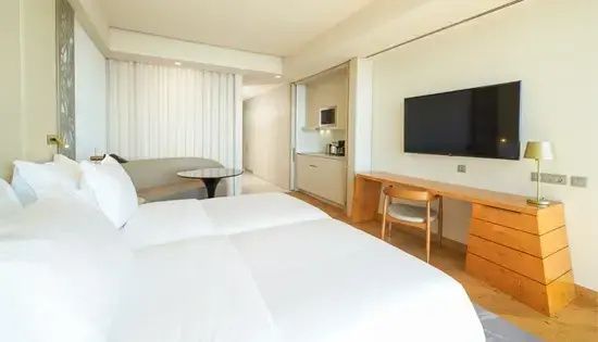 Helles Hotelzimmer mit Doppelbett, Fernseher an der Wand, Schreibtisch und integrierter Küchenzeile.