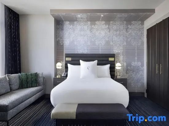 Zimmer mit großem Bett, beigen Kissen, gepolsterter Bank und verzierter Wand.