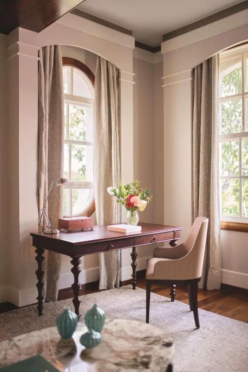 Eleganter Schreibtisch mit Blumenstrauß in einem Zimmer mit großen Fenstern.