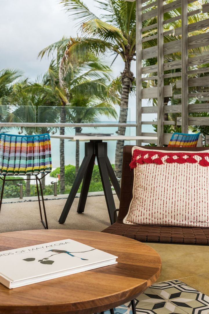 Terrasse mit bunten Stühlen, Holztisch und Kissen mit Blick auf Palmen und Meer.