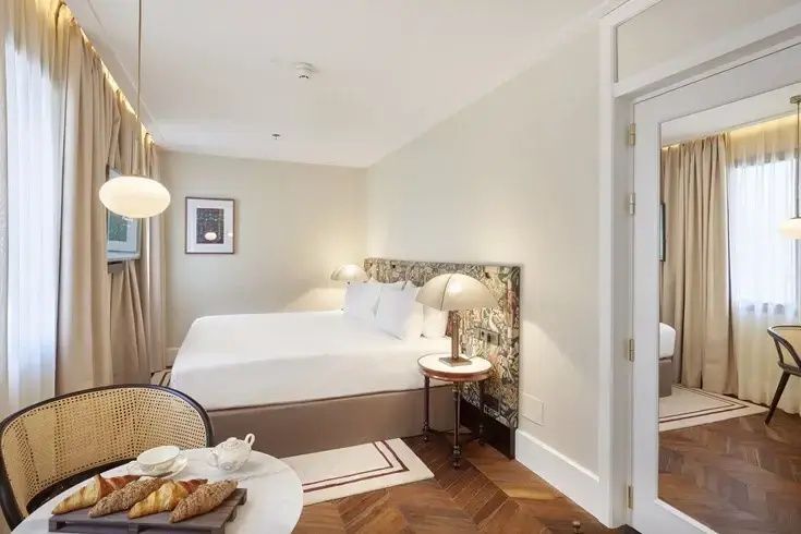 Helles Hotelzimmer mit Doppelbett, gemustertem Kopfteil, Parkettboden und einem kleinen Frühstückstisch.