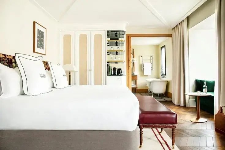 Helles Hotelzimmer mit weißem Doppelbett, roter Sitzbank und Blick ins Badezimmer.
