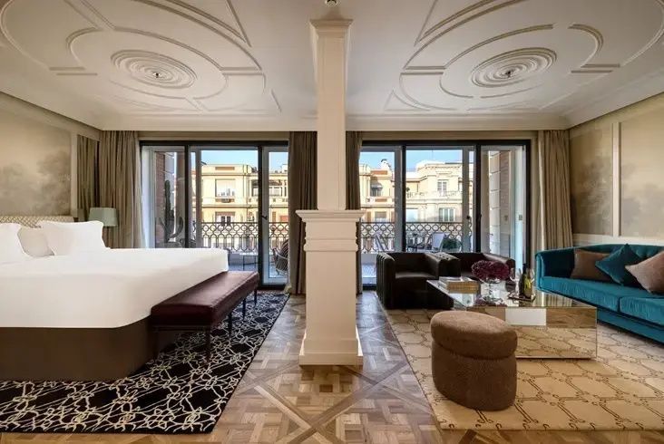 Suitenbereich mit Bett, Sitzgruppe und Fenstern mit Blick auf die Stadt.