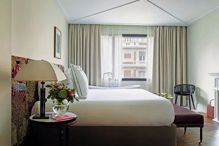 Doppelbett mit weißen Kissen, Nachttisch mit Blumenstrauß und Badewanne im Hintergrund.