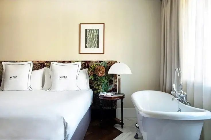 Bett mit besticktem Kopfteil, kleine Tischlampe und freistehende weiße Badewanne.