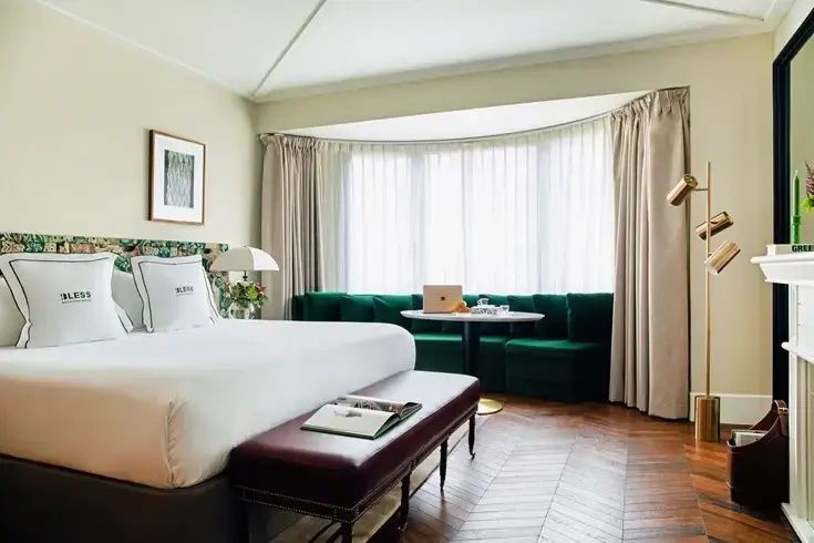 Helles Hotelzimmer mit Doppelbett, lederner Sitzbank und grüner Sofaecke am Erkerfenster.