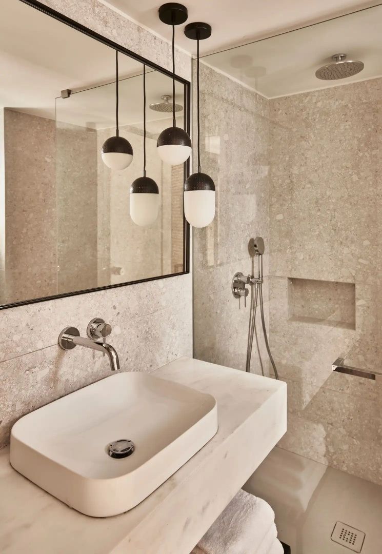 Badezimmer mit beigefarbenen Natursteinfliesen, Glasdusche und weißem Waschbecken.