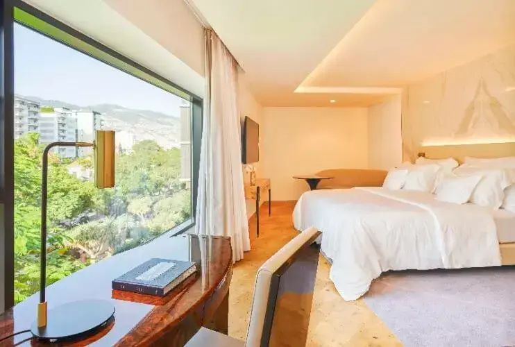 Helles Hotelzimmer mit großem Doppelbett, Schreibtisch und großem Fenster mit Blick auf Grünes.