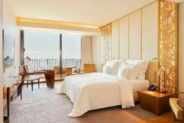 Helles Hotelzimmer mit weiß bezogenem Doppelbett und Balkontür mit Blick aufs Meer.