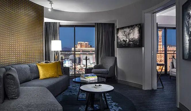 Wohnbereich eines Hotelzimmers mit grauem Sofa, Sessel und Fenster mit Stadtblick bei Dämmerung.