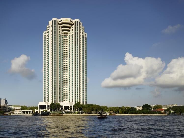 Hoch aufragendes, elegantes Gebäude des The Peninsula Bangkok direkt am Flussufer, umgeben von Bäumen und blauem Himmel mit wenigen Wolken.