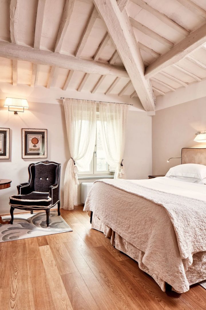 Schlafzimmer mit Holzbalkendecke, Doppelbett, dunklem Sessel und Fenstervorhängen.