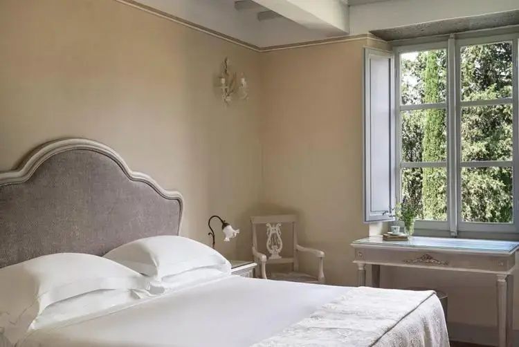 Schlafzimmer mit weiß bezogenem Bett, beigen Wänden und geöffnetem Fenster zum Garten.