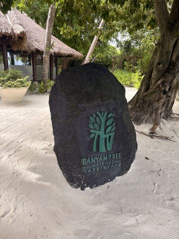 Steinerne Willkommenstafel mit dem Banyan-Tree-Logo im Sand unter einem alten Baum