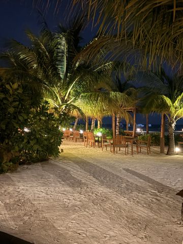 Holztische und Stühle im Sand unter beleuchteten Palmen am Abend, dahinter das Meer
