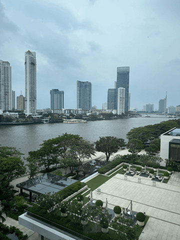 Durch die bodentiefen Fenster fällt der Blick auf den Chao Phraya River und die glitzernden Hochhäuser am gegenüberliegenden Ufer – eine eindrucksvolle Kulisse zwischen urbaner Energie und stiller Eleganz.