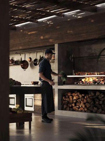 Koch an einem offenen Holzfeuergrill mit gestapeltem Kaminholz im Restaurant.