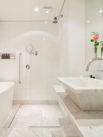Helles Badezimmer mit weißer Badewanne, gläserner Dusche und einem Waschtisch aus Marmor.