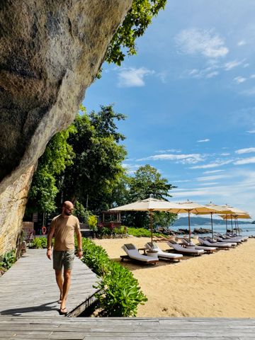 Mann spaziert entlang hölzernen Weges an tropischem Strand mit Liegen und Sonnenschirmen.