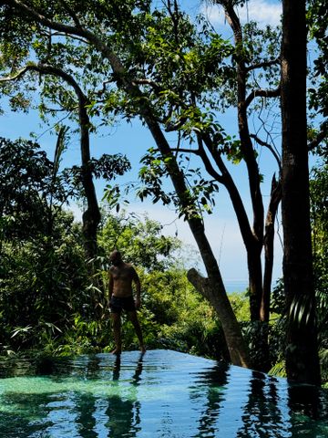 Mann am Infinity-Pool umgeben von tropischem Wald mit Meerblick im Hintergrund