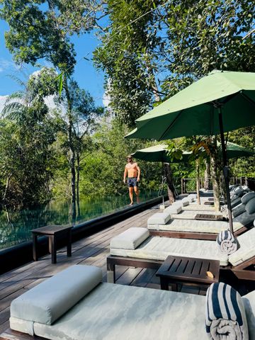 Elegante Liegestühle am Infinity-Pool umgeben von dichtem Grün. Einzelperson am Beckenrand.