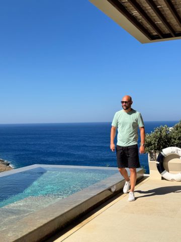 Mann auf Terrasse mit Meerblick und Infinity-Pool unter blauem Himmel.