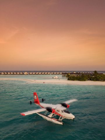 Wasserflugzeug über ruhigem Ozean vor einem sonnigen Resort bei Sonnenuntergang.