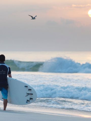 Surfer steht mit Brett am Strand und schaut auf die untergehende Sonne.