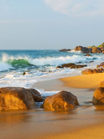 Wellen brechen gegen Felsen bei sonnigem Wetter am Strand.