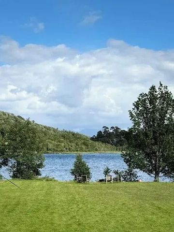 Grüne Wiese vor einem See mit bewaldeten Hügeln im Hintergrund.