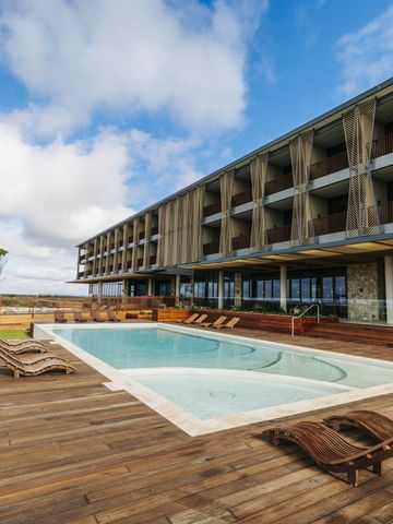 Pool mit Holzdeck und Liegen vor der Fassade des Resorts unter blauem Himmel