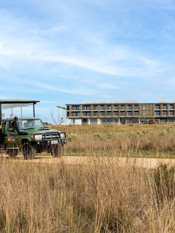 Offenes grünes Safarifahrzeug mit Gästen in der Graslandschaft, dahinter das Resortgebäude
