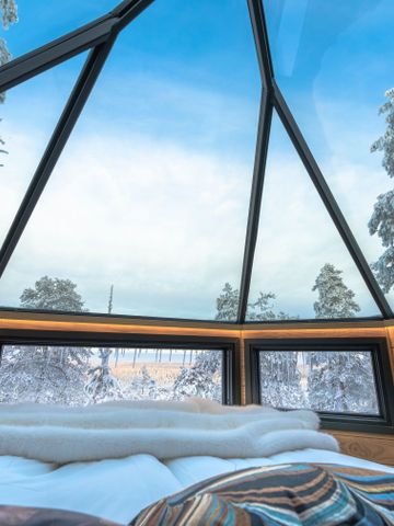 Bett unter dem Glasdach der Glass Igloo Suite, darüber verschneite Kiefern und blauer Himmel