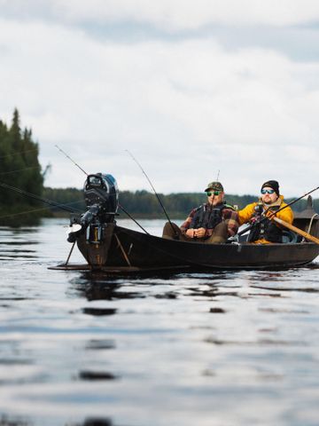 Zwei Angler im Holzboot beim Lachsfischen auf einem Fluss im Sommer, Ufer mit dichtem Wald