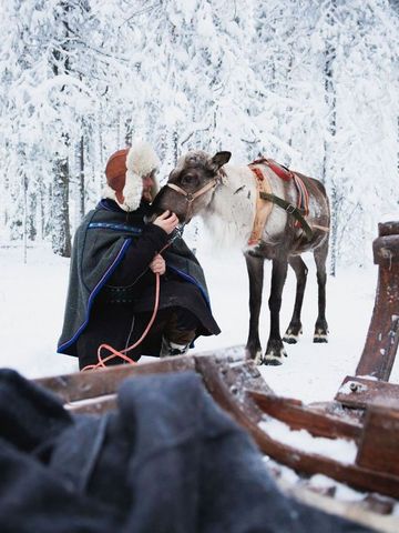 Rentierführer in traditioneller Kleidung mit Rentier und Schlitten im verschneiten Wald