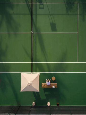Person sitzt unter Sonnenschirm auf einem Tennisplatz