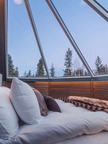 Doppelbett unter dem Glasdach eines Iglus mit Fellen, Kissen und Blick auf Kiefern im Abendlicht
