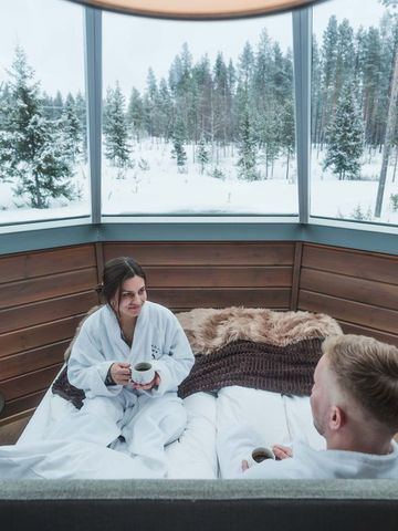 Paar im Bademantel mit Kaffee im Bett eines Glasiglus, durch die Fenster verschneiter Wald