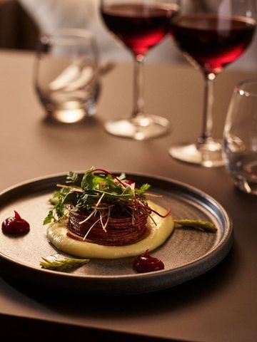 Teller mit Wildgericht, Püree und Kräutern auf dunklem Tisch, daneben zwei Gläser Rotwein