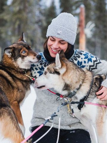 Frau im Strickpullover begrüßt zwei Huskys im Schnee vor dem Hundezwinger