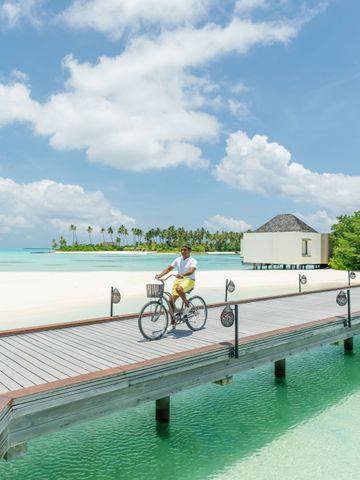 Mann fährt Fahrrad über Steg vor türkisfarbenem Wasser und weißem Sandstrand.