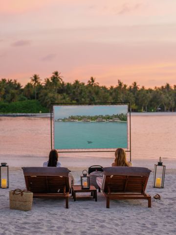 Zwei Personen auf Strandliegen betrachten eine Leinwand bei Sonnenuntergang am Strand.