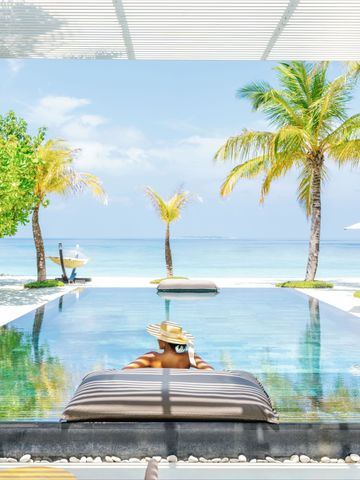 Person in Strohhut entspannt am Infinity-Pool mit Meerblick und Palmen.