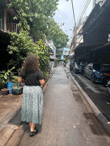 Mona ist von hinten zu sehen, wie sie mit der Kamera in der Hand durch die schmalen Gassen Bangkoks schlendert – umgeben von Streetart, kleinen Läden und urbanem Leben