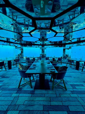 Unterwasserrestaurant mit eleganter Einrichtung, großen Fenstern und Blick auf die Meereswelt – umgeben von Fischen und türkisblauem Wasser.