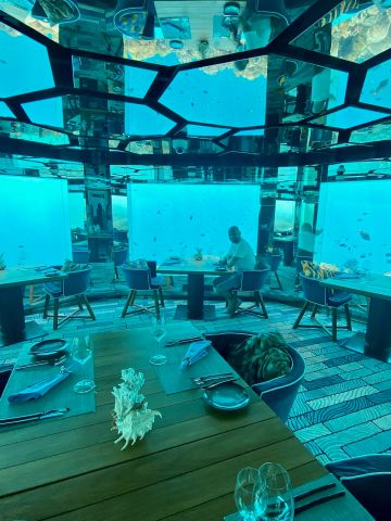 Blick in ein elegantes Unterwasserrestaurant mit gedeckten Tischen und Panoramafenstern, durch die Fische und Korallen im türkisblauen Meer zu sehen sind.