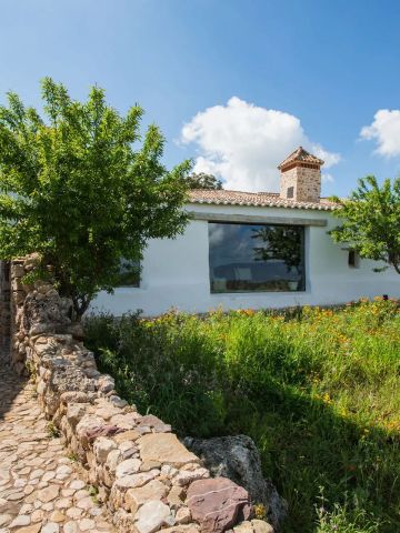 Außenansicht von La Donaira, einem Boutique-Hotel in Andalusien, mit weiß getünchtem Gebäude, Natursteinweg, blühender Wiese und ländlicher Idylle unter blauem Himmel.